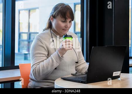 Femme engagée avec un ordinateur portable dans un espace de travail contemporain, tenant une tasse de café verte, entourée de lumière naturelle et d'éléments de design minimalistes Banque D'Images