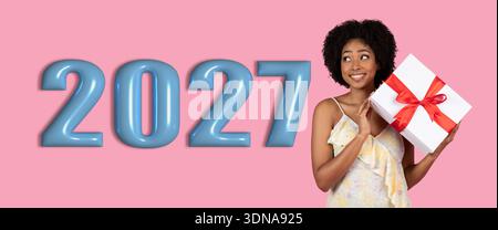 Jeune femme tient une boîte-cadeau à côté de Big Numbers célébrant l'année 2027 dans un cadre joyeux Banque D'Images