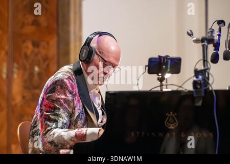 Homme âgé portant des écouteurs joue un piano à queue dans un cadre de performance intérieure, expression concentrée, veste colorée à motifs, microphones et re Banque D'Images