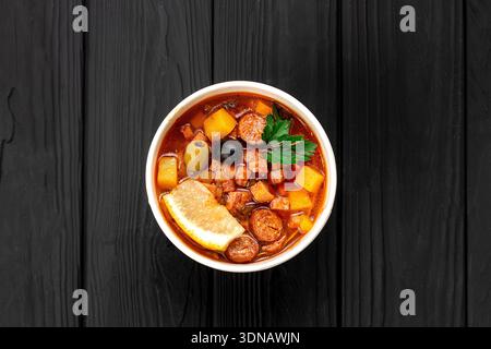 Soupe Solyanka avec saucisse, olives et citron dans un bol kraft avec de la crème sure sur une table sombre Banque D'Images