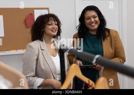 Diverses collègues féminines discutant des vêtements sur le rack avec des cintres dans la petite salle d'exposition tenant la tablette Banque D'Images