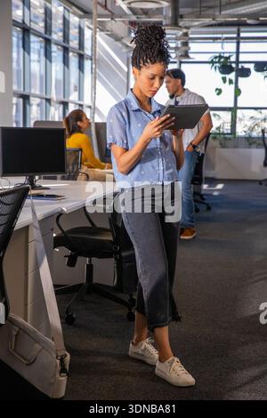 Divers collègues travaillant alors que femme afro-américaine tenant une tablette dans le bureau ouvert près des moniteurs Banque D'Images