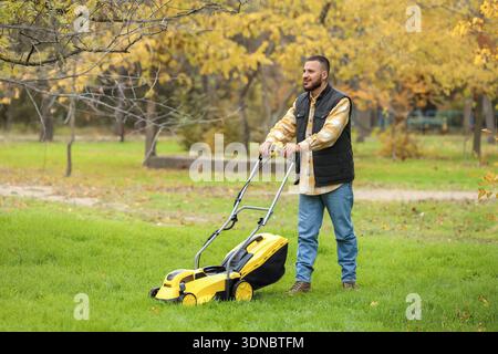 Beau jeune homme heureux taillant l'herbe avec une tondeuse à gazon moderne dans la cour arrière Banque D'Images