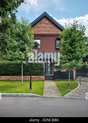 Maison moderne en briques rouges dans la verdure. Résidence de deux étages avec toit à pignons et accents métalliques noirs. Une pelouse verte et une passerelle pavée mènent à l'entrée. Banque D'Images