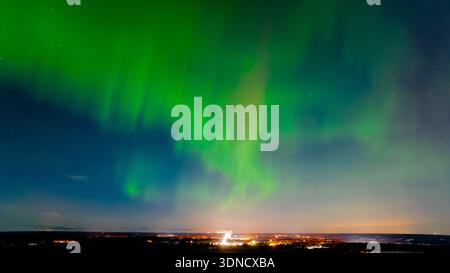 Vue aérienne des aurores boréales verdoyantes danses à travers le ciel sombre au-dessus des lumières scintillantes lointaines de la ville, Karis, Uusimaa, Finlande. Banque D'Images