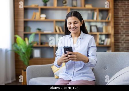 Jeune femme assise sur un canapé confortable dans son salon, se connectant joyeusement avec son téléphone portable pour la communication, les médias sociaux ou les activités en ligne, incarnant la connectivité moderne Banque D'Images