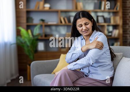Jeune femme souffrant de douleurs sévères à l'épaule et au bras, grimaçant et frottant son bras tout en étant assise sur un canapé dans un salon moderne, montrant inconfort et fatigue Banque D'Images