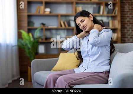 Jeune femme indienne assise sur un canapé massant le cou et les épaules, montrant des maux cervicaux et des tensions musculaires dues à une mauvaise posture, au stress et à la fatigue à la maison Banque D'Images