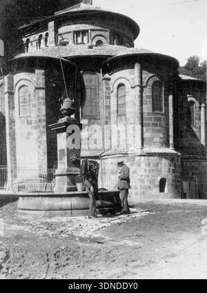 Cette photographie en noir et blanc capture l'abside romane de l'église de la Gorgon à Orcival, en France. Deux hommes en tenue du début du XXe siècle se tiennent près d'une fontaine de pierre au premier plan. La maçonnerie massive en pierre, les fenêtres cintrées et la conception circulaire à plusieurs étages de l'église soulignent son style architectural Auvergne historique sous un ciel clair. D'une collection de photographies uniques du XXe siècle. Photographe amateur, clichés et quelques-uns plus grands dans un seul album, retraçant les voyages européens d'une famille. Banque D'Images