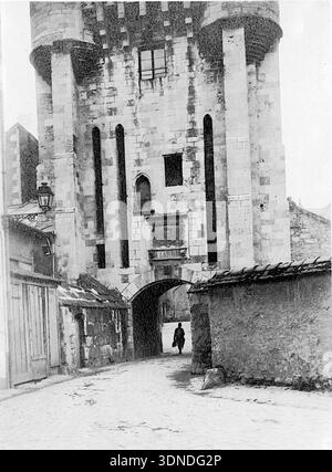 Une grande tour de pierre s'élève au-dessus de la rue pavée, sa passerelle voûtée formant un passage sous d'étroites ouvertures verticales et des détails sculptés. Des bâtiments adjacents altérés avec des toits de tuiles encadrent l'approche, tandis qu'une figure solitaire marche à travers l'arche. La scène reflète le caractère architectural du début du siècle de Nevers. D'une collection de photographies uniques du XXe siècle. Photographe amateur, clichés et quelques-uns plus grands dans un seul album, retraçant les voyages européens d'une famille. Banque D'Images