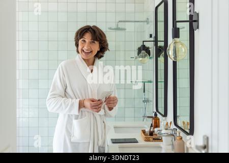 Femme d'âge moyen portant un peignoir éponge blanc debout à la vanité tenant la soie dentaire près de la pomme de douche à effet pluie Banque D'Images