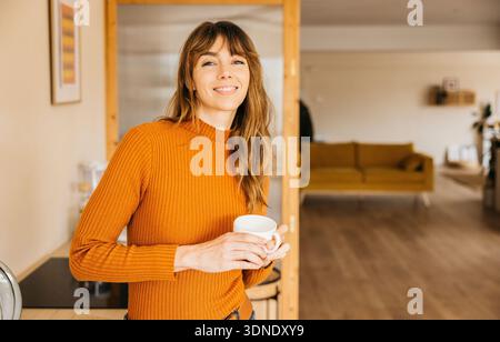 Femme souriant tout en tenant une boisson chaude, profitant d'une matinée domestique à la maison Banque D'Images