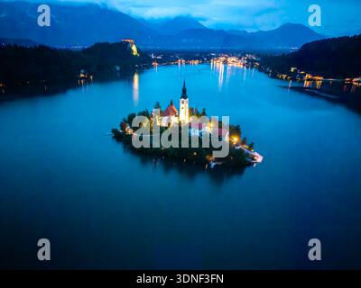 Vue aérienne de l'église illuminée de Marie la Reine sur l'île de Bled au milieu des eaux bleues sombres et sereines, avec le château de Bled perché au sommet d'une lointaine salut Banque D'Images