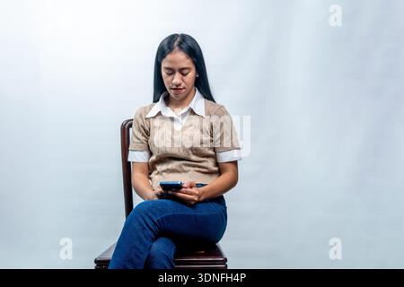 Une jeune femme indonésienne assise sur une chaise, regardant son téléphone portable avec une expression concentrée sur un fond blanc de studio. Banque D'Images