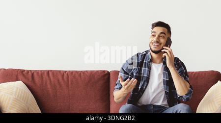 Homme assis sur le canapé faisant un appel téléphonique tout en souriant et en parlant dans une salle lumineuse Banque D'Images