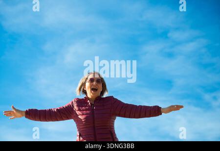 Femme heureuse avec des lunettes de soleil debout avec les bras grands ouverts contre un ciel bleu. Concept de liberté, de joie et de fitness en plein air Banque D'Images