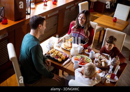 Une famille se réunit dans un restaurant pour prendre le petit déjeuner. Ils partagent une table remplie de nourriture. Les parents parlent avec leurs deux enfants, créant un joyeux a Banque D'Images