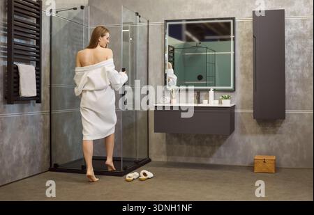 Vue arrière d'une femme portant un peignoir blanc, allant avoir une douche dans la cabine de douche Banque D'Images