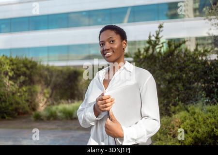 Jeune femme d'affaires confiante en chemise blanche avec ordinateur portable debout près de l'immeuble de bureaux moderne. Professionnelle réfléchie souriant et regardant loin Banque D'Images