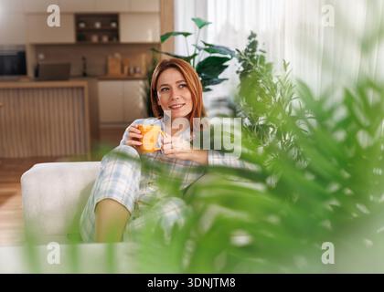 Une femme est assise sur un canapé en pyjama et tient un verre tout en regardant par la fenêtre. Des plantes vertes l'entourent dans l'espace du salon. Cuisine avec Banque D'Images