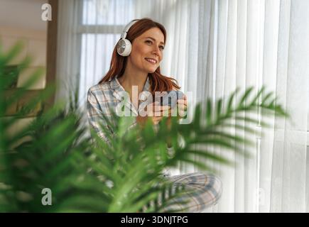 Une femme en pyjama est assise près d'une fenêtre portant des écouteurs et sourit en utilisant son téléphone. Elle aime la musique dans un espace intérieur confortable avec des plantes à proximité. Banque D'Images