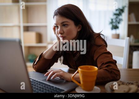 Une femme est assise à une table dans un salon. Elle se concentre sur son ordinateur portable tout en sirotant du thé. Elle se sent fatiguée et s'ennuie Banque D'Images