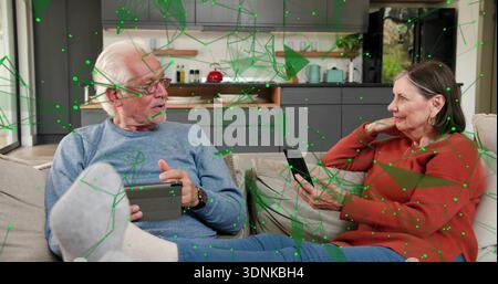 Assis couple senior dans des chandails à l'aide d'une tablette et d'un smartphone dans le salon avec maillage numérique Banque D'Images