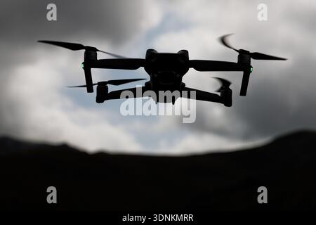 Un drone en silhouette vole contre un ciel nuageux sombre avec des formes de montagne en dessous. Banque D'Images
