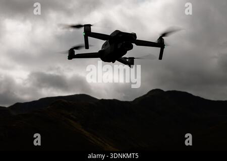 Un drone en silhouette vole contre un ciel nuageux sombre avec des formes de montagne en dessous. Banque D'Images