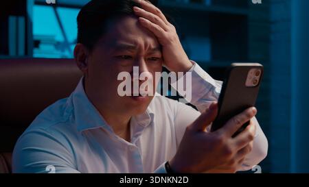 Bouleversé souligné l'échec asiatique d'homme d'affaires d'âge moyen avec téléphone mobile la nuit bureau soir triste frustré homme d'affaires coréen avec smartphone échouer Banque D'Images