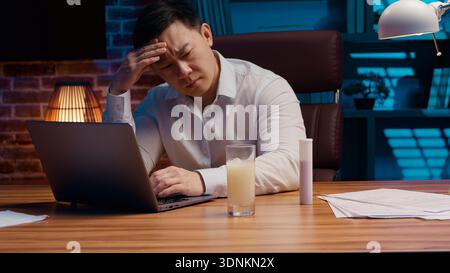 Homme d'affaires asiatique malade avec ordinateur portable jetant la pilule dans le verre d'eau homme d'affaires coréen sentir mal de tête trouble du sommeil insomnie surmené mâle japonais Banque D'Images