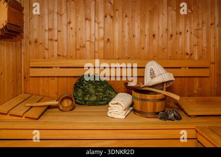 Seau en bois avec cuillère, chapeau de sauna, serviettes et balai sur banc. Détails intérieurs et accessoires traditionnels du sauna finlandais Banque D'Images