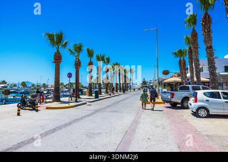 Belle vue sur la rue bordée de palmiers avec des touristes marchant le long de la promenade du port près de la marina et des cafés en bord de mer. Kardamena. Grèce. Banque D'Images