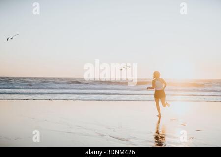 Femme courant sur la plage au coucher du soleil avec des mouettes en vol et du sable humide réfléchissant Banque D'Images
