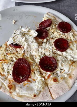 Pizza blanche avec fromage fondu et saucisse épicée tranchée servie sur une assiette, partiellement consommée. Ambiance décontractée mettant en valeur des plats simples et réconfortants Banque D'Images