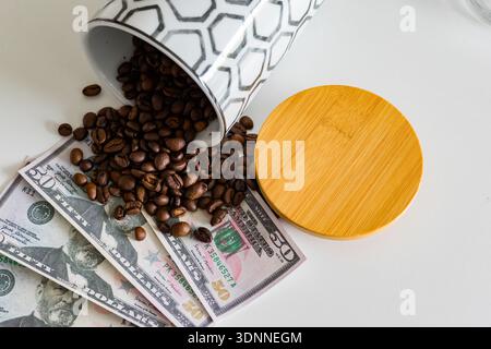 Une photo conceptuelle en grand angle de grains de café torréfiés foncés débordant d'un récipient en céramique sur plusieurs billets de 50 dollars américains. Un couvercle en bambou repose Banque D'Images