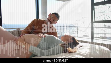 Partenaire souriant penché sur la femme enceinte en haut vert, en plaçant la main sur le ventre sur le lit, superpositions Banque D'Images