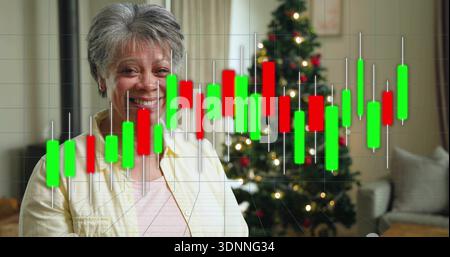 Femme âgée souriante en chemise rose-beige assise à la maison avec arbre de Noël et tableau de chandelier Banque D'Images
