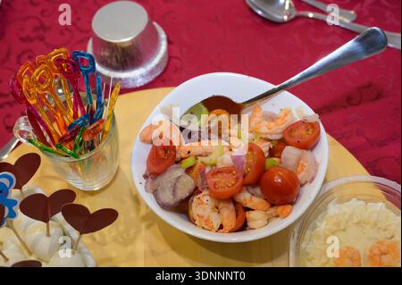 Un bol de salade de crevettes et de tomates repose sur une table avec une cuillère et une tasse. Le bol est rempli de crevettes, tomates et oignons, et il y en a plusieurs Banque D'Images