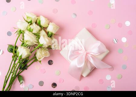 Bouquet de roses blanches avec boîte cadeau et ruban rose sur fond pastel avec des confettis. Vue de dessus, pose à plat. Banque D'Images