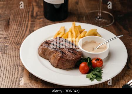 Succulent steak de bœuf accompagné d'une sauce crémeuse au poivre et de frites croquantes, garni de tomates cerises et de persil frais, servi dans une assiette blanche Banque D'Images