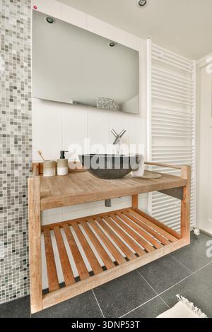 La salle de bains moderne dispose d'une élégante coiffeuse en bois avec un élégant lavabo noir. Les textures contrastées du dosseret carrelé et du sol brillant cre Banque D'Images