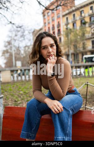 Jeune femme aux cheveux ondulés est assise sur un banc rouge, portant un haut marron et un Jean. Elle regarde attentivement la caméra, posant son menton sur sa main. URB Banque D'Images