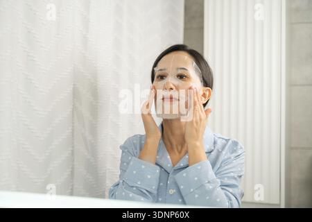 Une femme portant un pyjama applique soigneusement un masque facial dans sa maison. Elle se tient devant un miroir, s'assurant que le masque s'adapte correctement à optima Banque D'Images