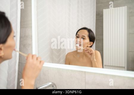 Une femme se tient devant un miroir de salle de bain se brossant les dents, pratiquant une bonne hygiène bucco-dentaire. Son reflet est visible dans le miroir, montrant un clea Banque D'Images