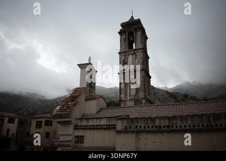 Tour d'église historique et toits de tuiles à Speloncato, Corse, encadrés par des sommets de montagne brumeux sous ciel couvert. Banque D'Images