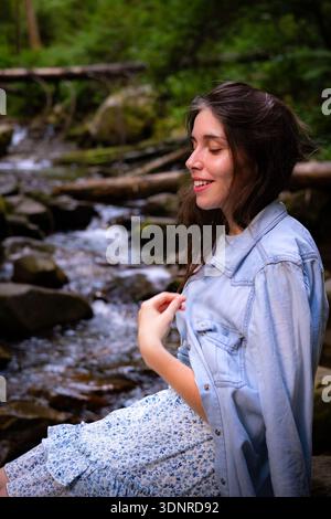Une jeune femme se détendant au bord d'un ruisseau forestier, profitant d'un moment paisible entouré d'une nature verdoyante. La lumière naturelle douce, la faible profondeur de champ, Banque D'Images