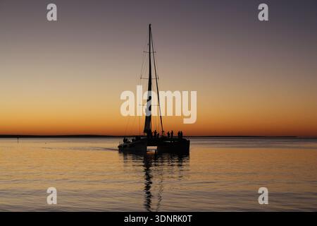 Silhouette d'un voilier au coucher du soleil sur l'eau calme avec ciel orange et reflet Banque D'Images