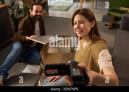 Jeune femme adulte caucasienne enregistrant une vidéo avec un caméscope tout en souriant, assise sur le sol à côté d'un homme d'âge moyen caucasien assemblant des meubles, une boîte en carton et des outils visibles Banque D'Images