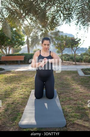 Une femme enceinte en vêtements sportifs noirs s'agenouille sur un tapis de yoga dans un parc ensoleillé, vérifiant sa montre intelligente. La scène transmet le fitness, le bien-être de la maternité Banque D'Images